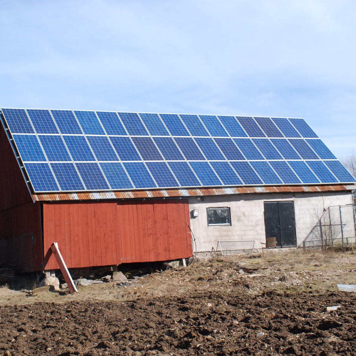 Solpaneler installerade på taket av en röd ladugårdsliknande struktur på ett öppet fält under en klar himmel.