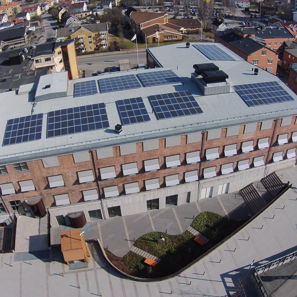 Flygfoto över en tegelbyggnad med solpaneler på taket, omgiven av urbana strukturer.