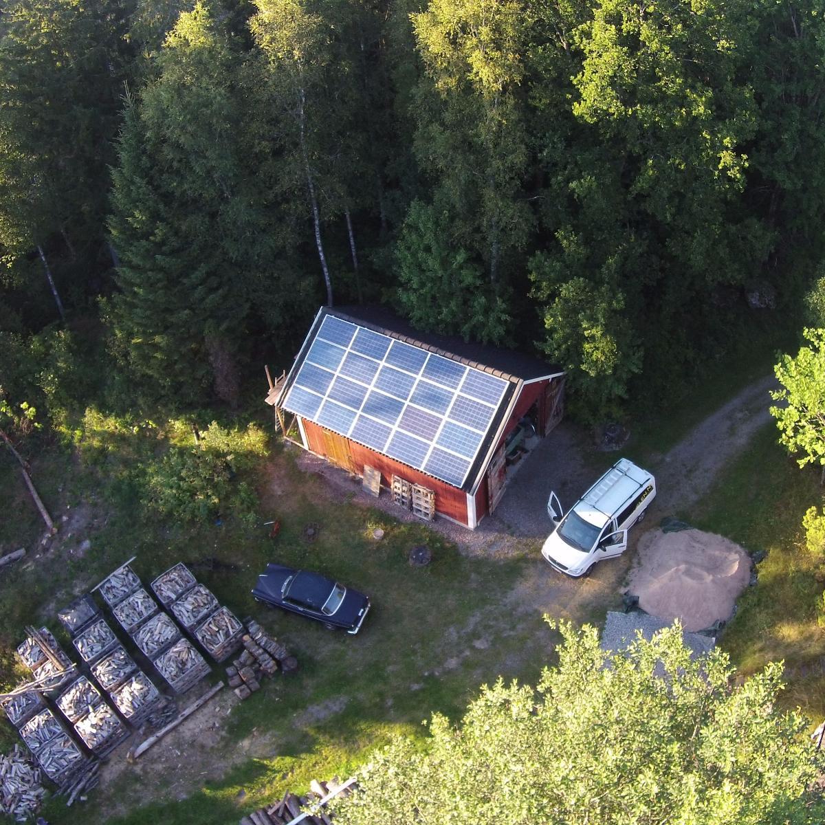 Flygfoto över en stuga med solpaneler på taket, omgiven av träd. Två fordon står parkerade i närheten, med vedhögar synliga till vänster.