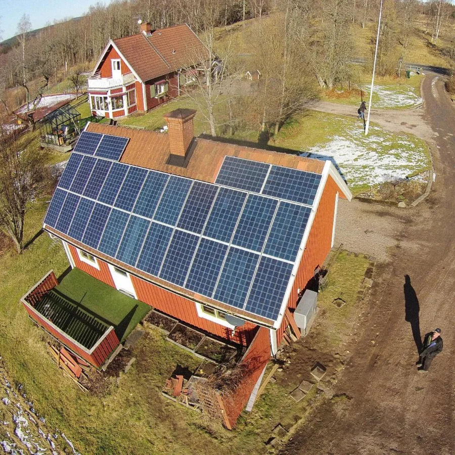 Rött hus med solpaneler på taket, omgivet av ett lantligt landskap med en väg, kala träd och fläckig snö. En person går i närheten.
