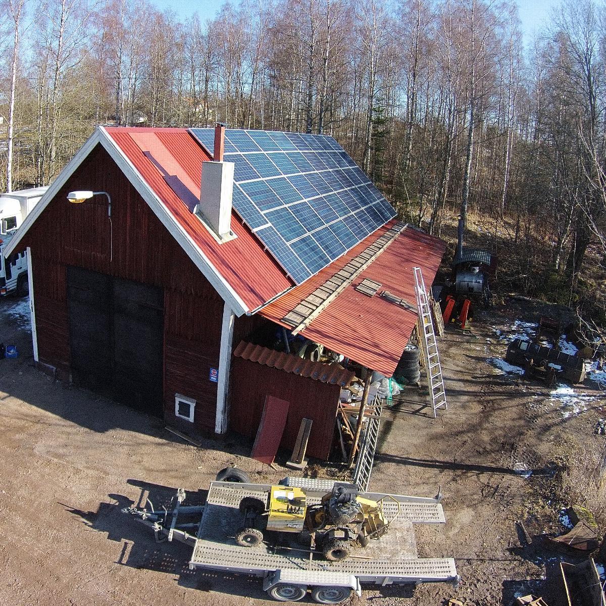 En lantlig byggnad med rött tak har solpaneler ovanpå. Det omgivande området inkluderar stegar, maskiner och utspridda föremål. Träd och en smutsmark är synliga.