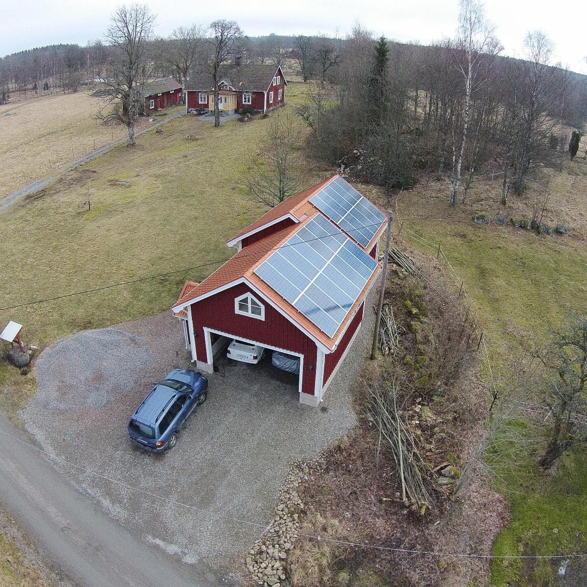 Flygfoto över en röd lada med solpaneler på taket. En blå bil står parkerad på grusuppfarten. Runt ladugården finns träd och gräsfält. Ett hus syns i bakgrunden.