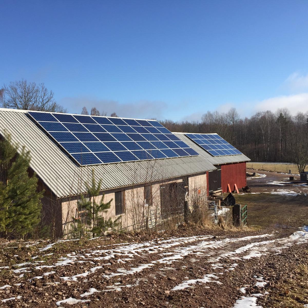 Lantlig scen med solpaneler på ladugårdstak under en klarblå himmel. Glesa snöfläckar täcker den omgivande marken och lövlösa träd finns i bakgrunden.
