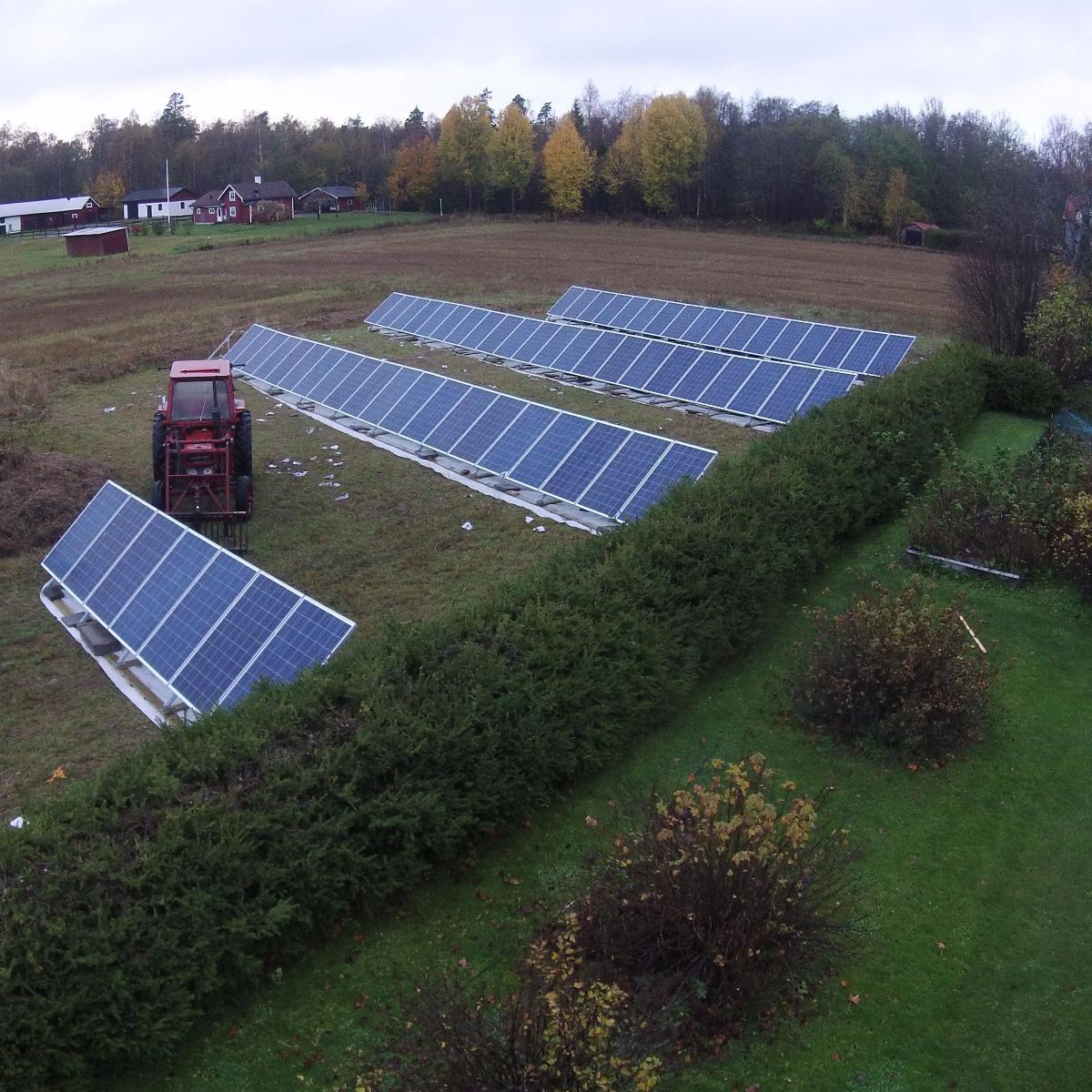 Flygfoto över flera rader av solpaneler i ett fält, kantat av gröna häckar, med en närliggande röd traktor och omgivande träd.