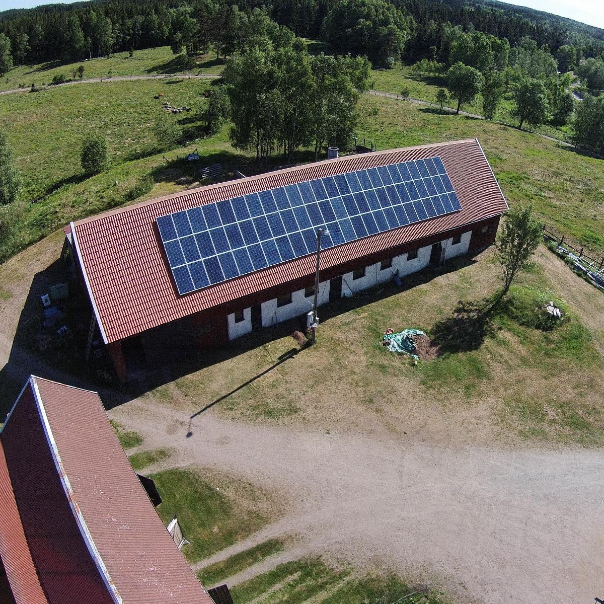 Flygfoto över en lång rektangulär byggnad med solpaneler på taket, omgiven av gräsbevuxna fält och träd.