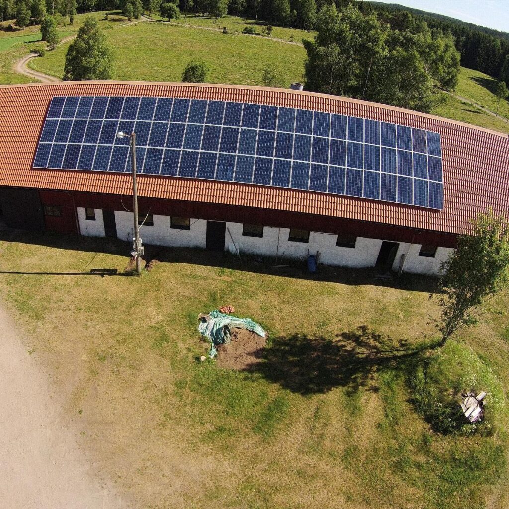 Flygfoto över en byggnad med solpaneler på ett rött tegeltak, omgivet av gräs och träd.
