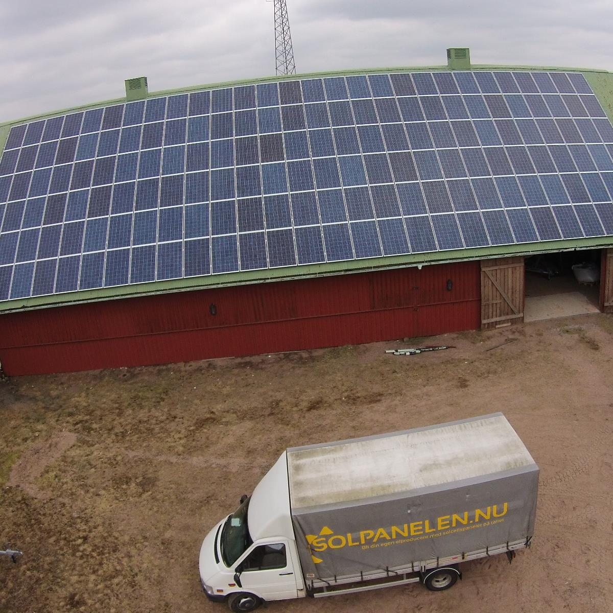 En vit lastbil med texten SOLPANELEN.NU står parkerad framför en röd lada med ett stort utbud av solpaneler på taket.