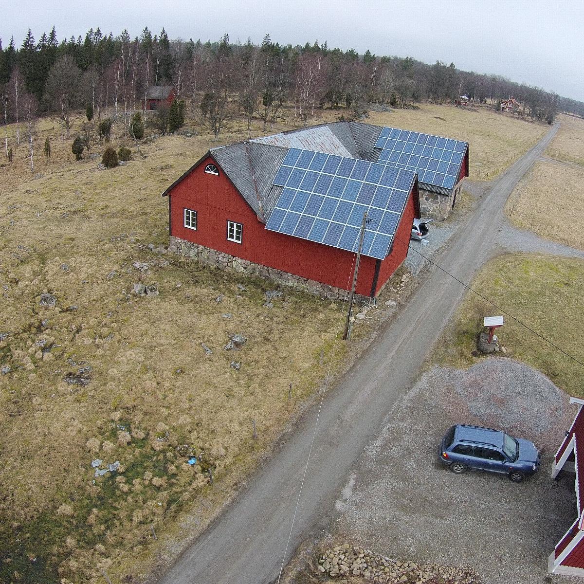 Flygfoto över en röd ladugård med solpaneler på taket, omgiven av gräs och träd, belägen intill en grusväg. En parkerad bil syns nära ladugården.