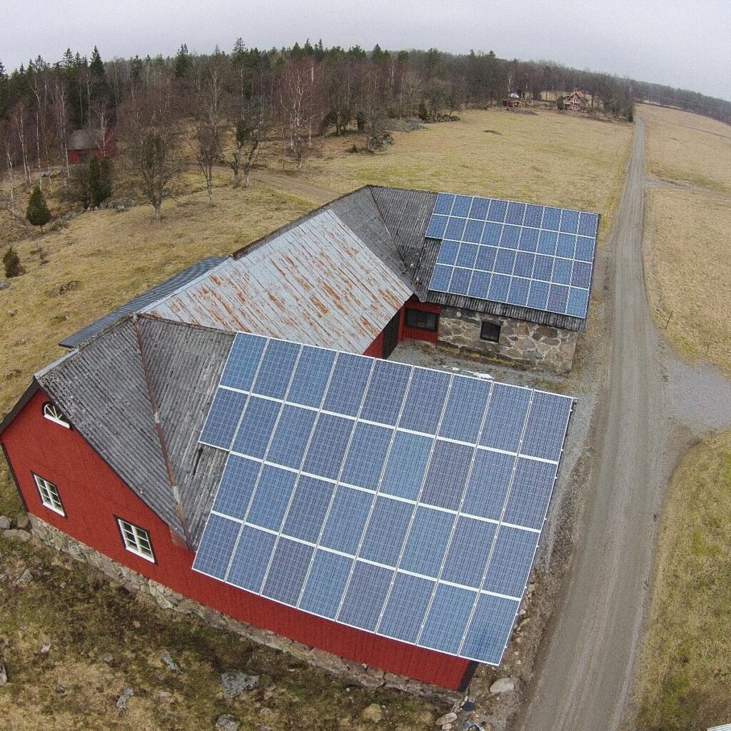 Flygfoto över en träbyggnad med solpaneler på taket, omgiven av gräs och träd, med en grusväg som leder bort.