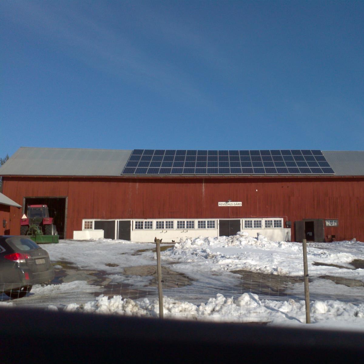 En röd ladugård med solpaneler på taket, omgiven av snö. Ett fordon är parkerat i närheten.