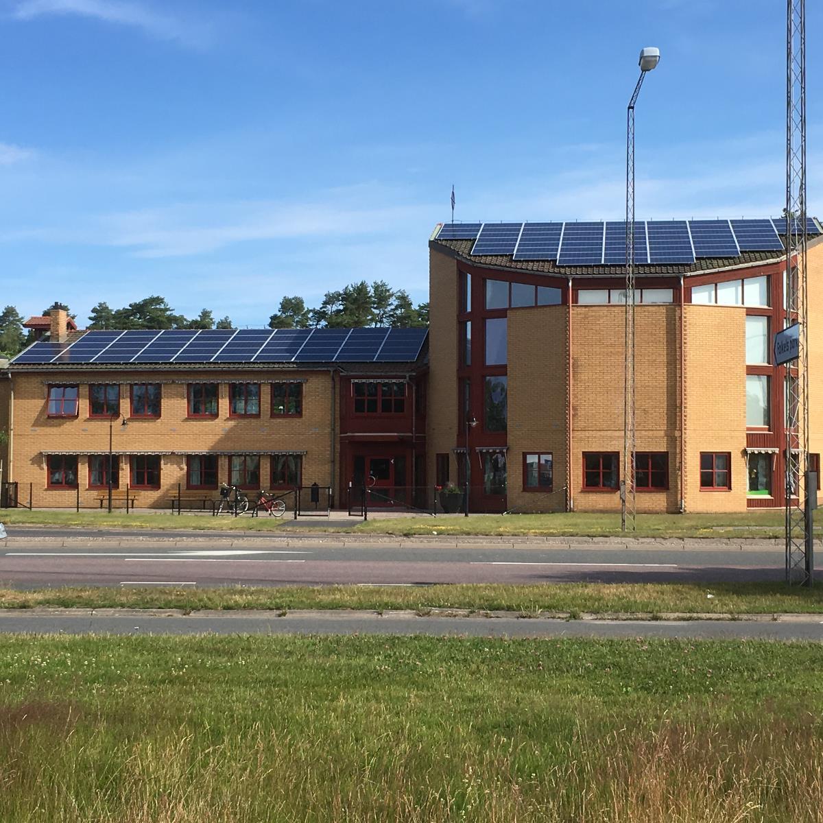 En tegelbyggnad med solpaneler på taket, belägen bredvid en väg med cyklar parkerade framför, under en klarblå himmel.