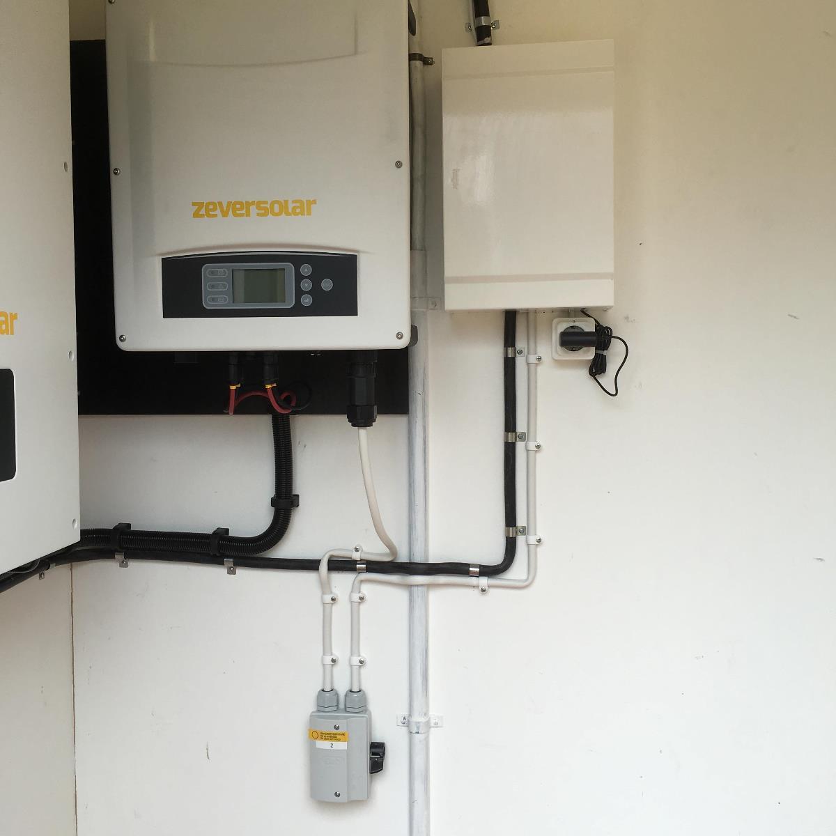 En Zeversolar inverter monteras på en vägg med anslutna elkablar. I närheten finns en vit elbox installerad ovanför ett uttag.