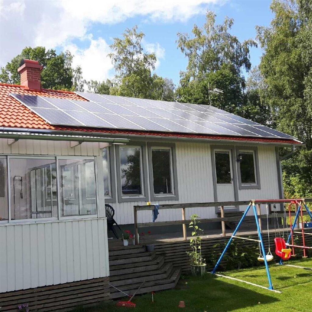 Ett vitt hus med rött tak har rader av solpaneler. En gungställning och en gräsbevuxen gård finns i förgrunden. Träd omger huset under en delvis molnig himmel.