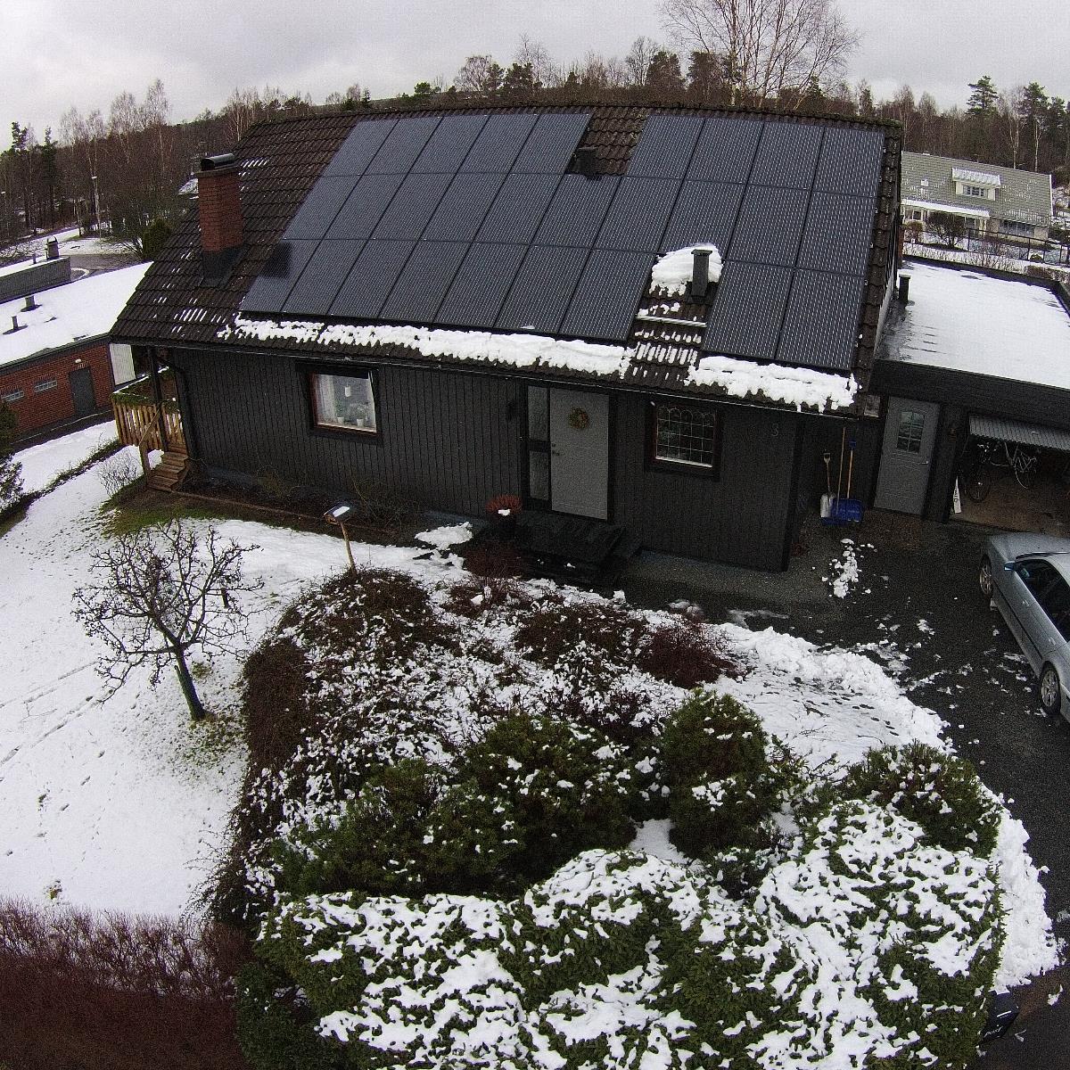 Hus med snötäckt tak, med solpaneler och en sektion utan paneler. Omgivande gård delvis täckt med snö. Mulen himmel.