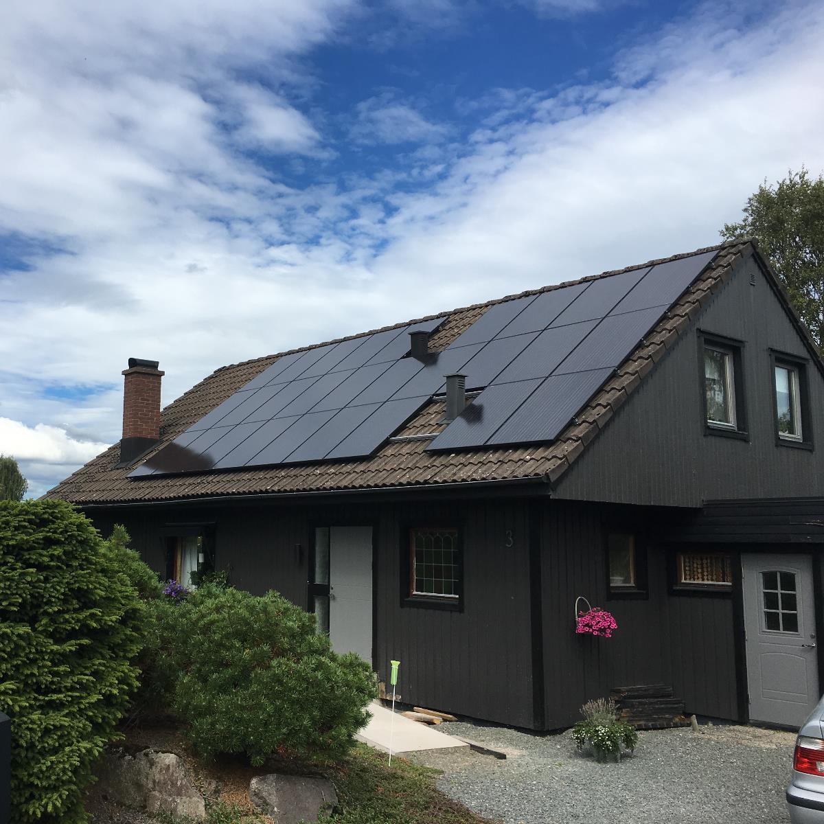 Mörkt trähus med solpaneler på taket, omgivet av buskar och en klar himmel.
