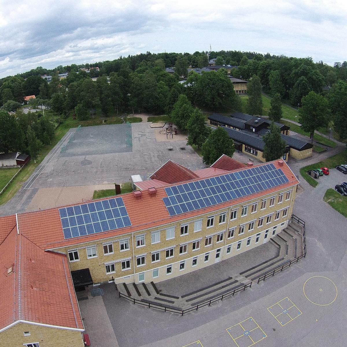 En flygvy över en skolbyggnad med solpaneler på taket, omgiven av träd och i anslutning till en lekplats och parkeringsplats.