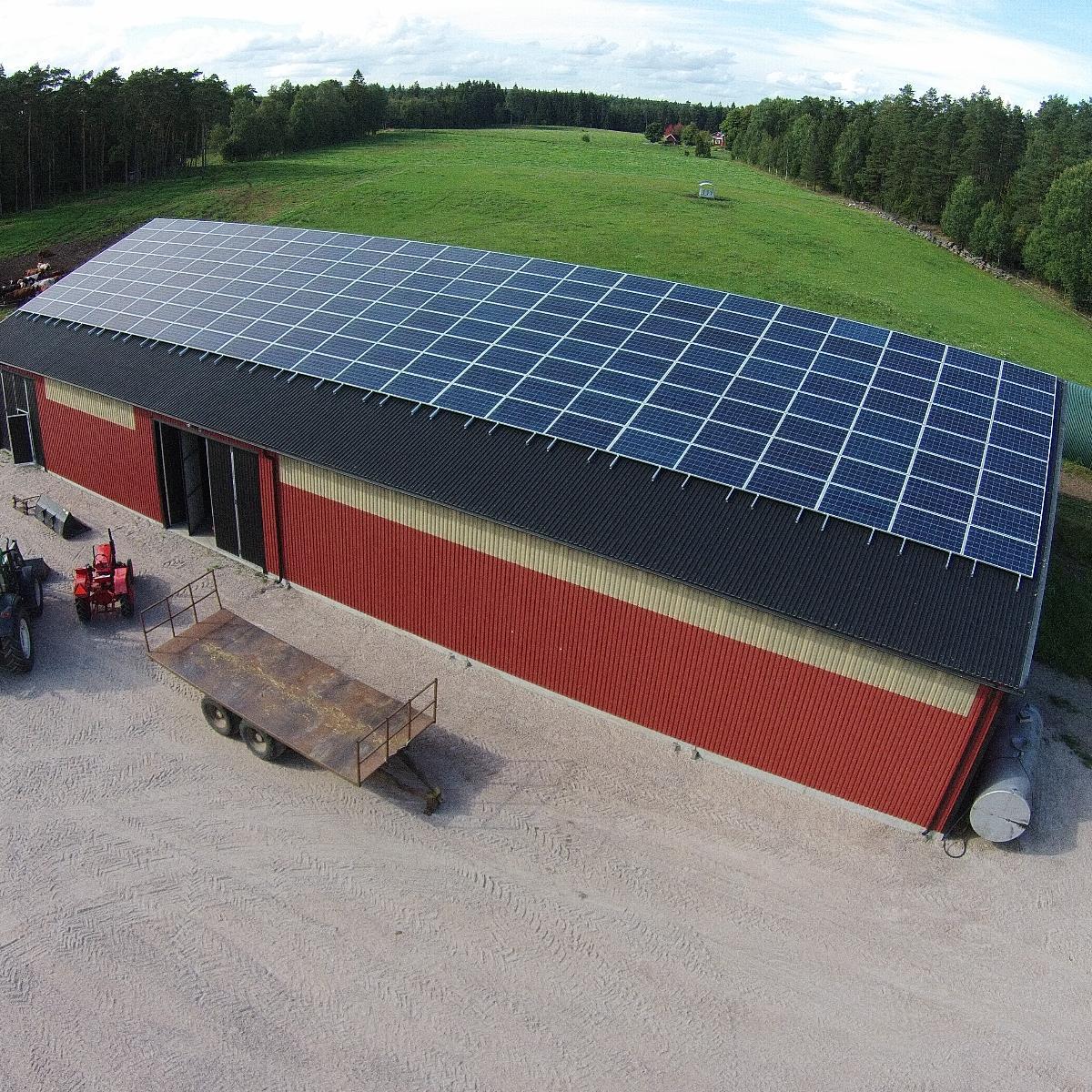 En stor, röd ladugård med plåttak täckt av solpaneler ligger på landsbygden omgiven av gröna fält och träd. En röd traktor och en flakvagn står parkerade i närheten.