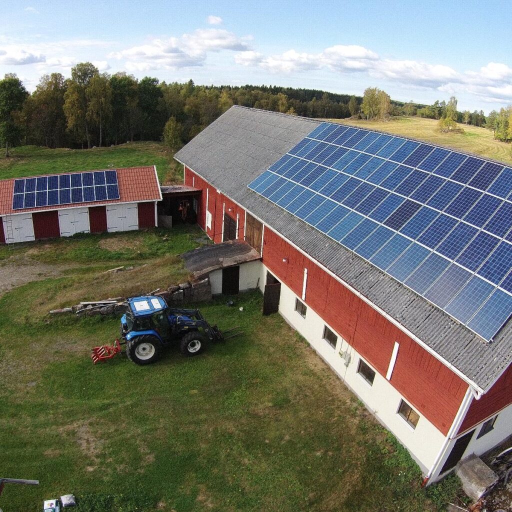 Röda lador med solpaneler på taken, omgivna av träd och åkrar. En blå traktor står parkerad framför den större ladugården. Himlen är delvis molnig.