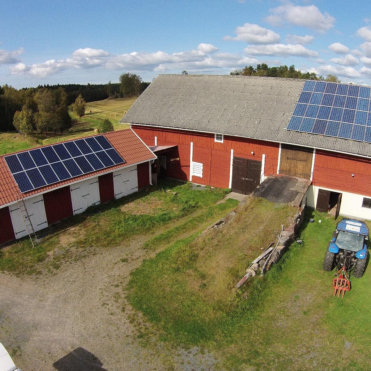 En gård med röda byggnader har solpaneler på hustaken. En blå traktor står parkerad i närheten. Åker och träd är i bakgrunden under en delvis molnig himmel.