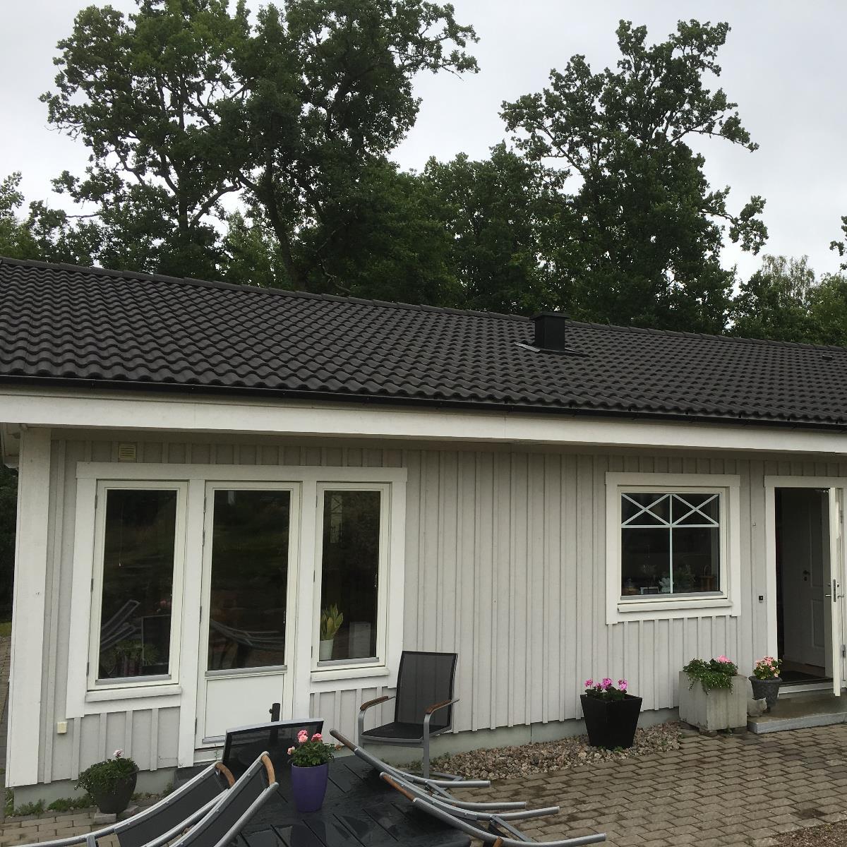 Litet enplanshus med ljusgrå träväggar, mörkt tak och vita inramade fönster. Utestolar och krukväxter står på en uteplats i förgrunden. Träden är i bakgrunden.