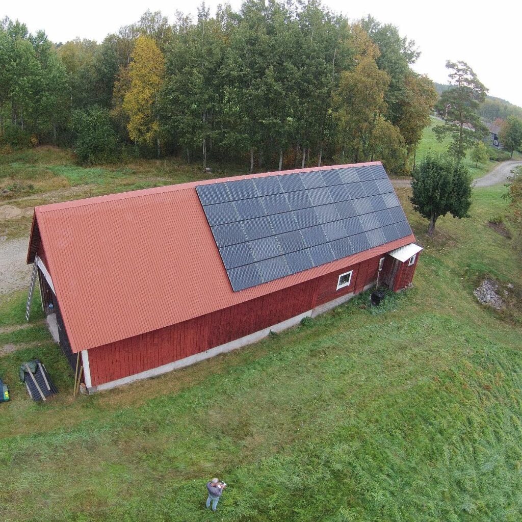 Röd ladugård med solpaneler på taket, omgiven av gräs och träd. En person står i gräset och tar ett foto.