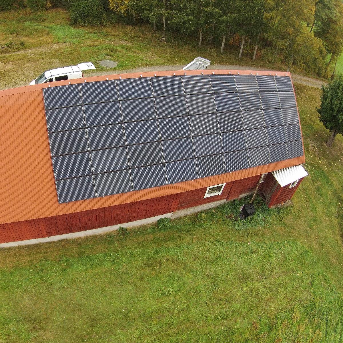 Flygfoto över en röd byggnad med en stor solpanel på taket, omgiven av gräsbevuxen mark och träd.