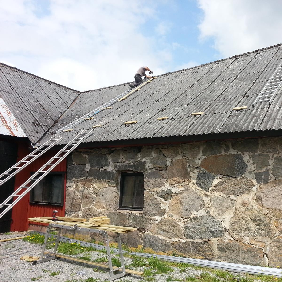 Person som reparerar ett sluttande korrugerat tak på en stenbyggnad med hjälp av stegar. Flera träplankor och verktyg ligger på marken. Himlen är delvis molnig.