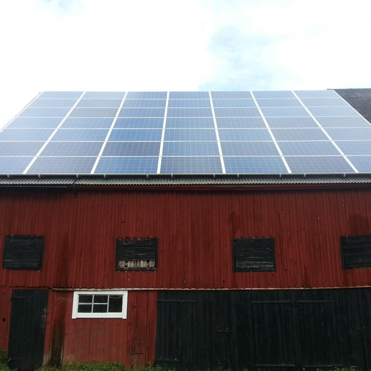 Stora solpaneler installerade på taket av en röd lada med mörka luckor, under en molnig himmel.