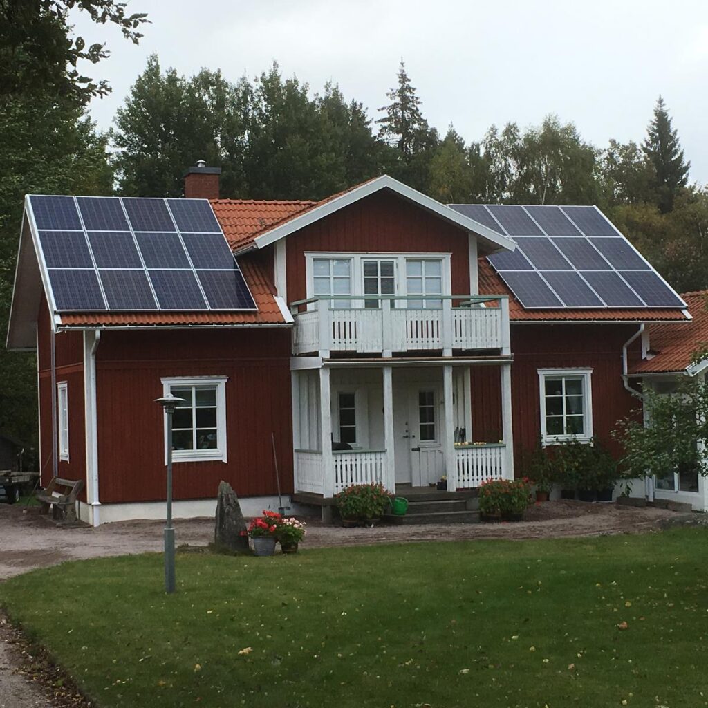 Rött hus med solpaneler på taket, vita fönster och trim, omgivet av träd och gräs, molnig himmel.