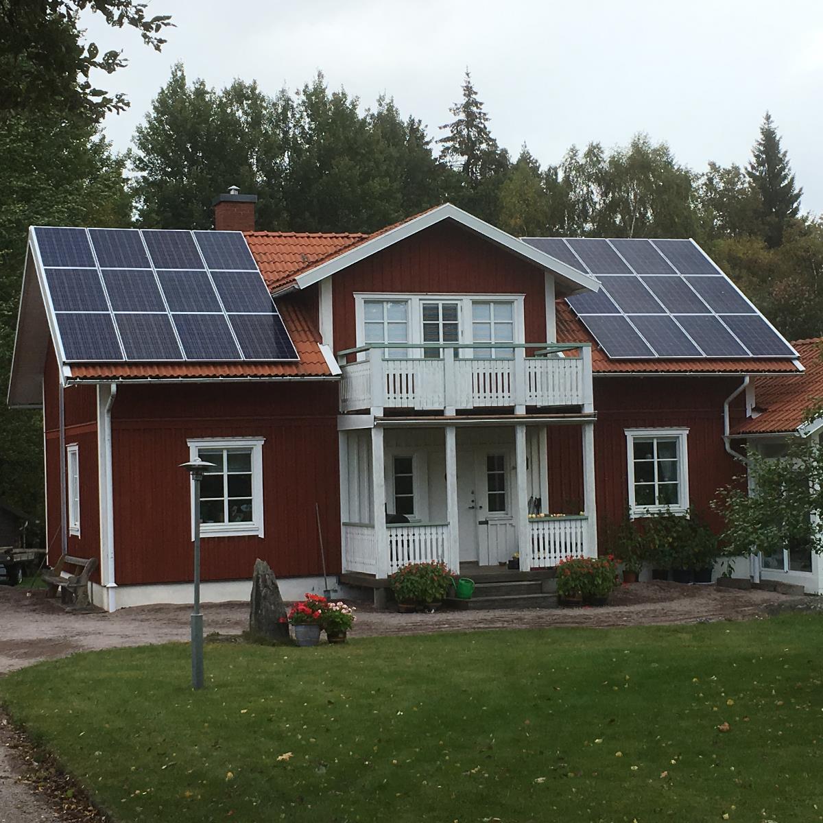 Rött hus med solpaneler på taket, vita fönster och trim, omgivet av träd och gräs, molnig himmel.