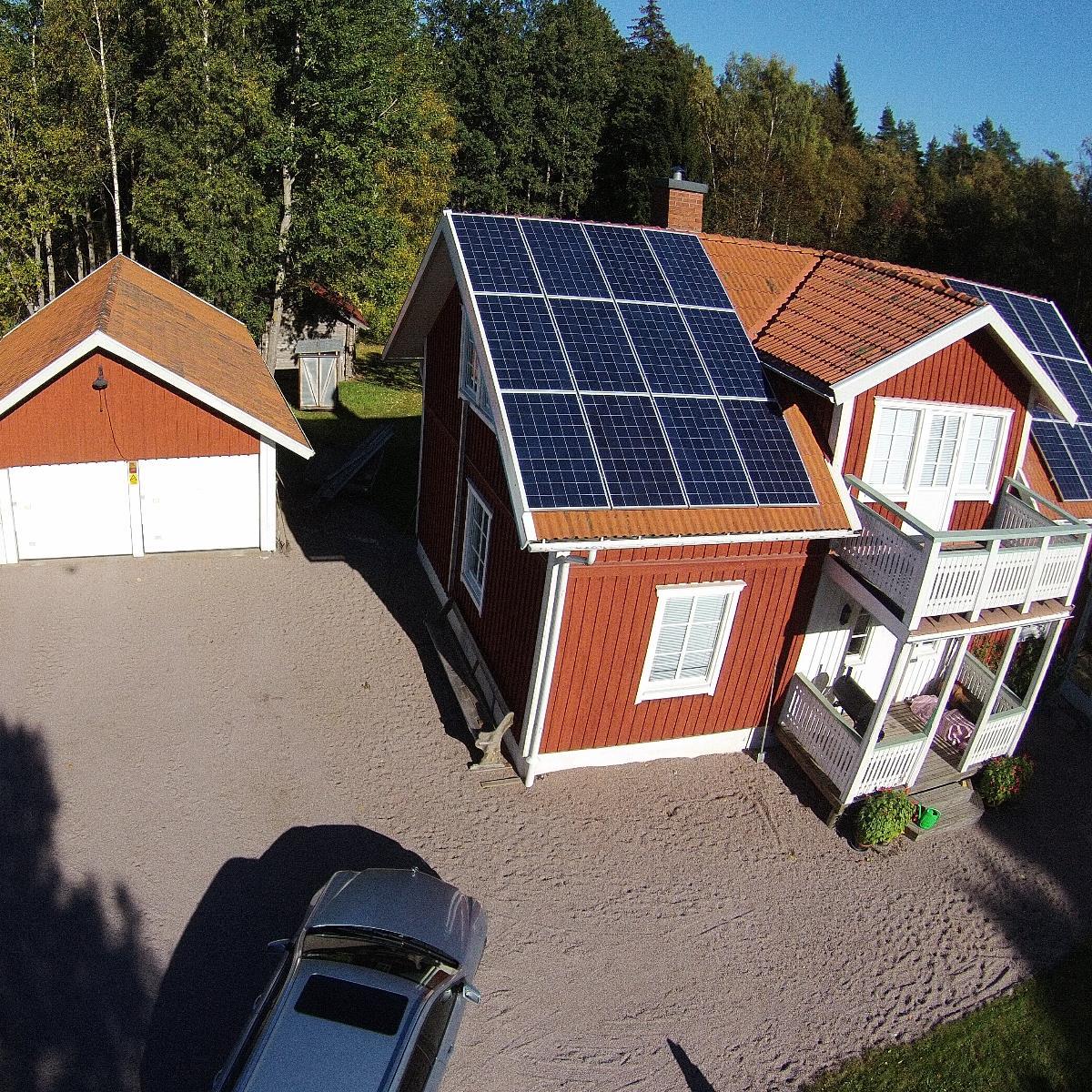 Ett rött trähus med solpaneler på taket, bredvid ett matchande garage, omgivet av träd. En parkerad bil syns på uppfarten.