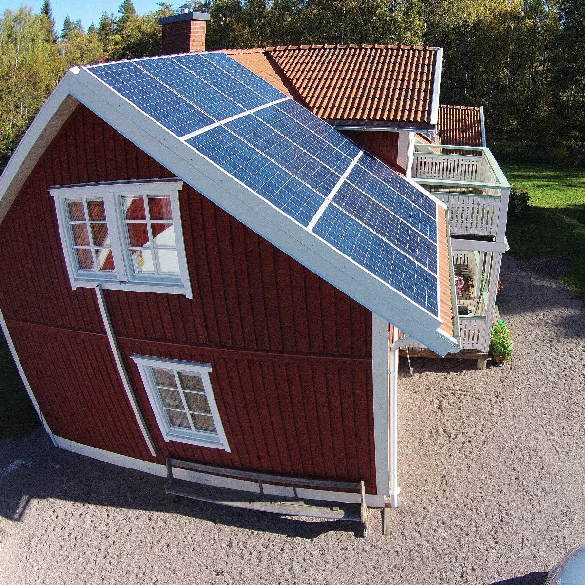 Ett rött hus med sluttande tak är försett med solpaneler. Det finns en balkong med vita räcken och träden syns i bakgrunden.