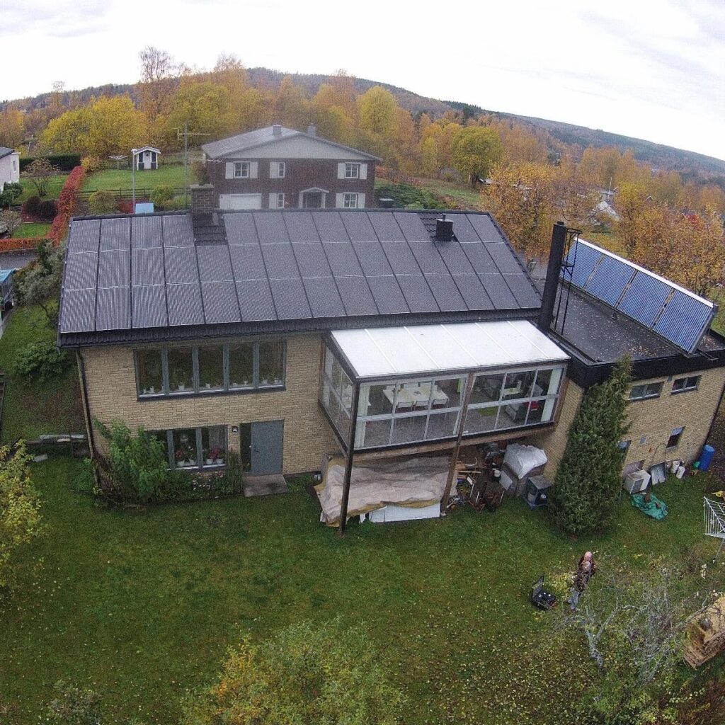 Flygfoto över ett tvåvåningshus med solpaneler på taket, omgivet av gräs och träd med höstlövverk. En inglasad veranda är synlig på baksidan.