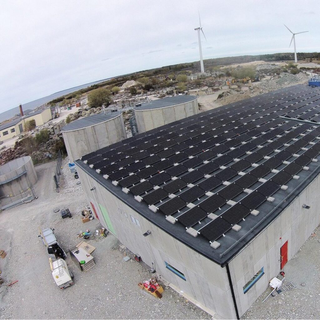 Taksolpaneler på en industribyggnad, med vindkraftverk och stenig terräng i bakgrunden.