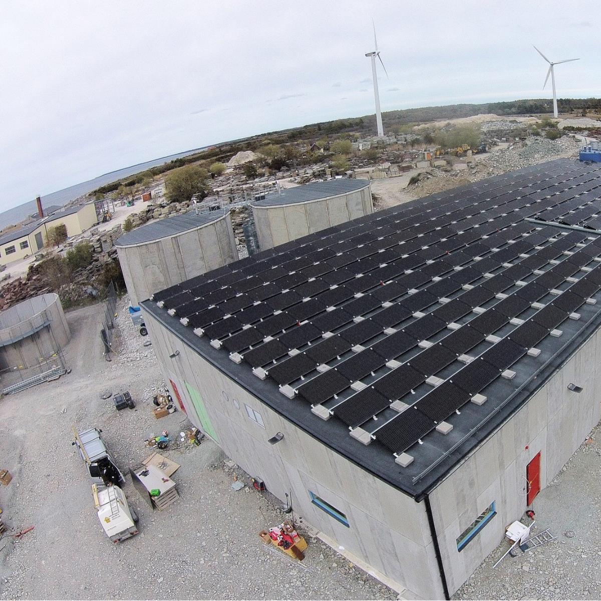 Taksolpaneler på en industribyggnad, med vindkraftverk och stenig terräng i bakgrunden.