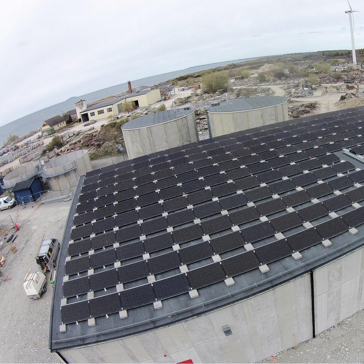 Takvy av en stor industribyggnad täckt med solpaneler. Omgivande terräng är industriell med strukturer och ett vindkraftverk synligt i bakgrunden.