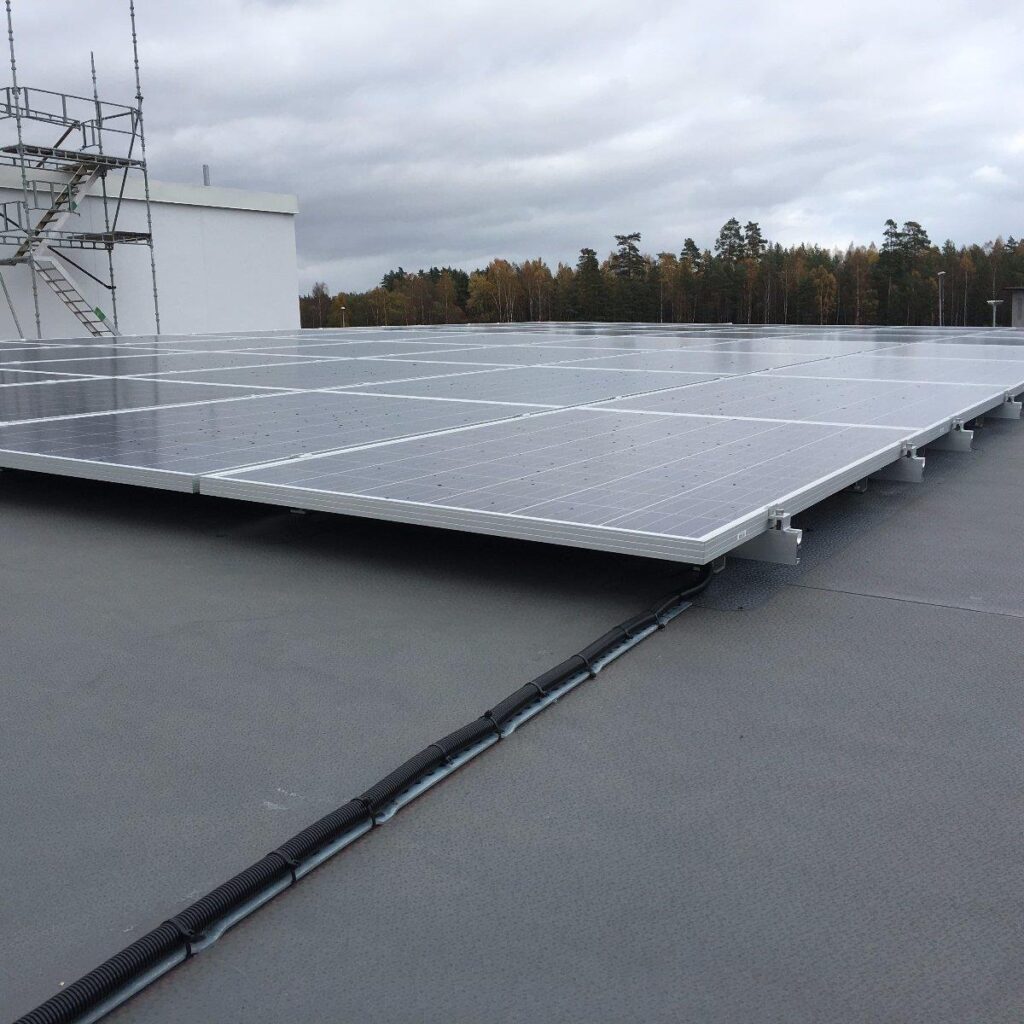 Solpaneler installerade på ett platt tak, med byggnadsställningar i närheten och en skog i bakgrunden under en molnig himmel.