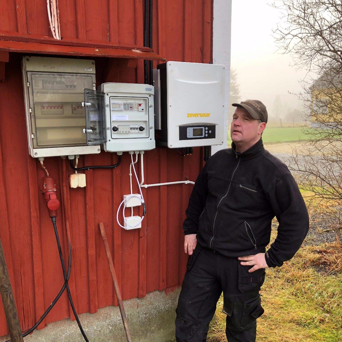 Person i arbetskläder står bredvid elektrisk utrustning monterad på en röd trävägg utomhus.