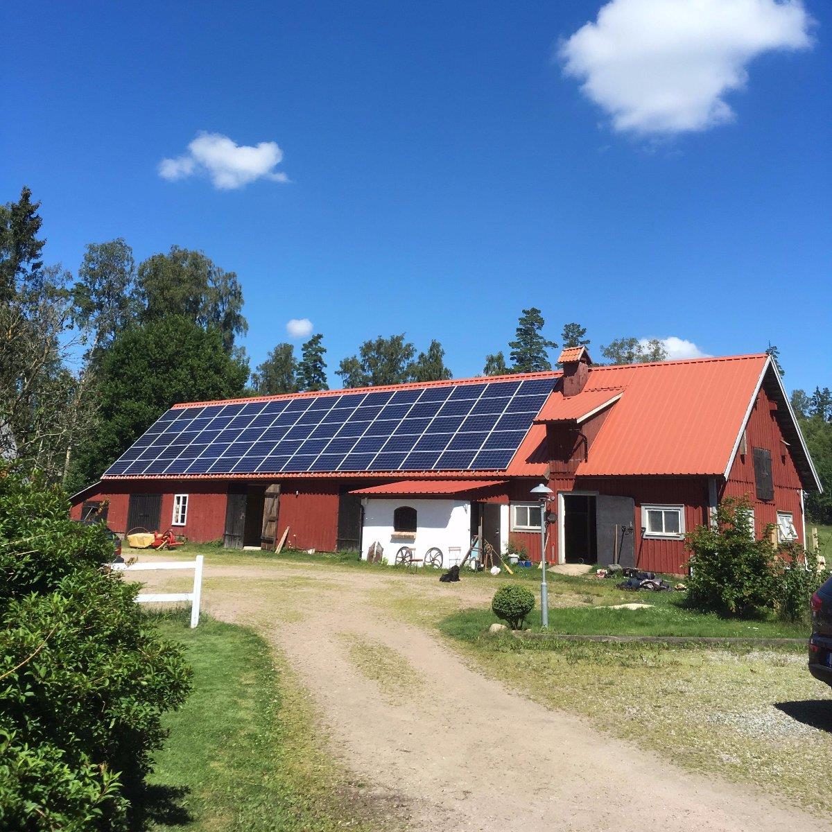 En röd ladugård med ett rött tak täckt av solpaneler, i ett lantligt landskap med träd och en klarblå himmel. En grusuppfart leder upp till ladugården.