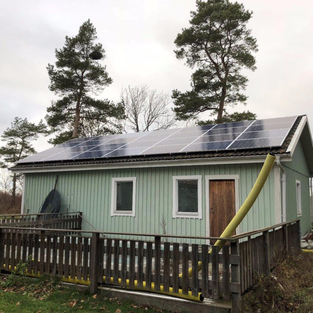 Litet grönt hus med solpaneler på taket, omgivet av ett trästaket. Två höga träd är i bakgrunden. Ett gult rör är fäst vid huset.