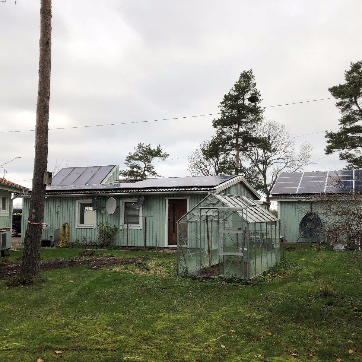 Ett litet grönt hus med solpaneler på taket och ett växthus i trädgården, omgivet av gräs och träd en molnig dag.