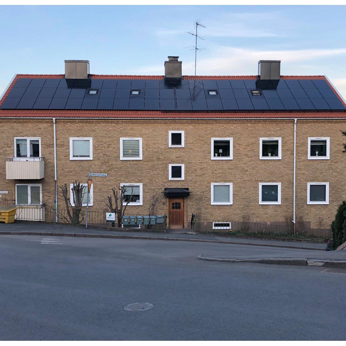 En tvåvånings tegelbyggnad med solpaneler på taket, flera fönster och en central trädörr. Gatan framför har en trottoar och en avfallscontainer.