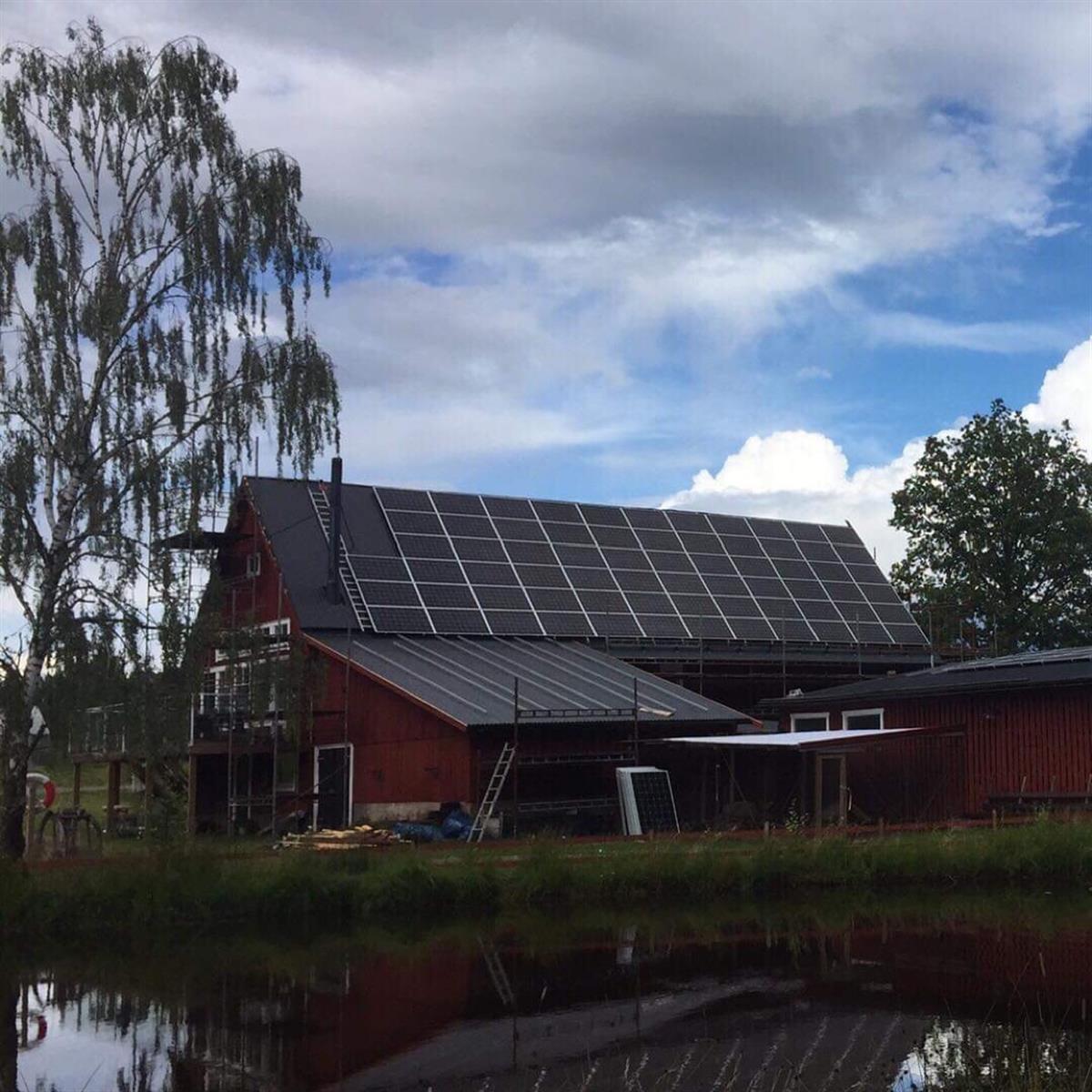 En röd ladugårdsliknande byggnad med solpaneler på taket reflekteras i en närliggande damm, med ett träd till vänster och en halvmolnig himmel ovanför.
