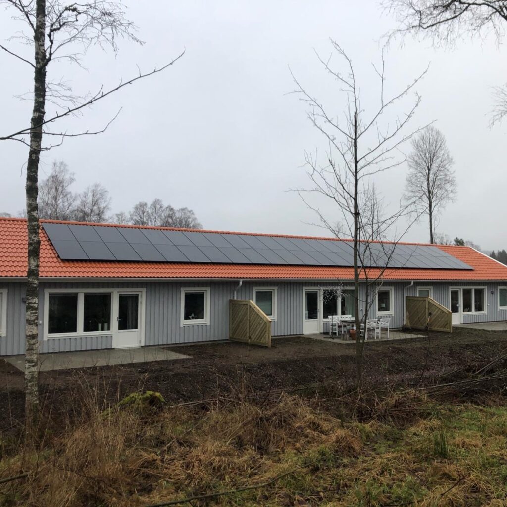 Envåningsbyggnad med rött tegeltak och solpaneler, omgiven av kala träd och gräs, under en molnig himmel.