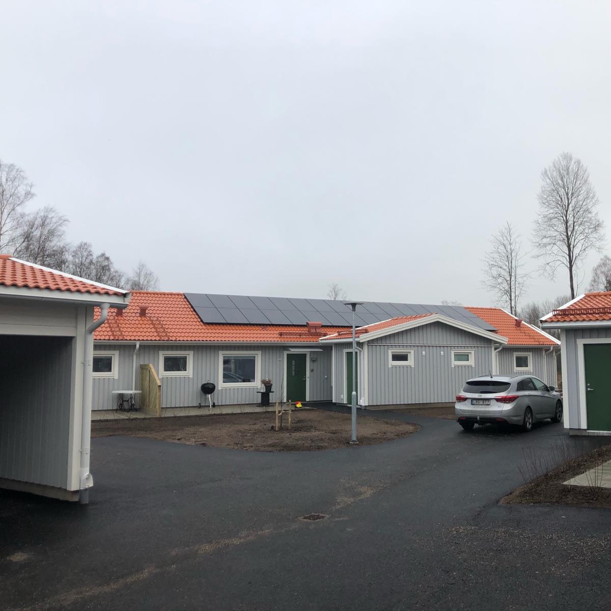 Enplanshus med rött tegeltak och solpaneler, mulen himmel. Vita sidospår, parkerad bil på uppfarten, täckt parkering till vänster, kala träd i bakgrunden.