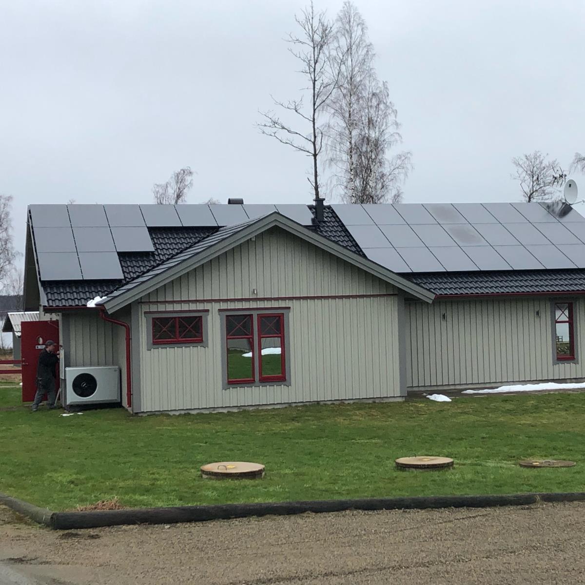Hus med solpaneler på taket, ljusgrå träbeklädnad, röd trim och omgivande grönt gräs. En person står nära en utomhusenhet på vänster sida. Mulen himmel ovanför.