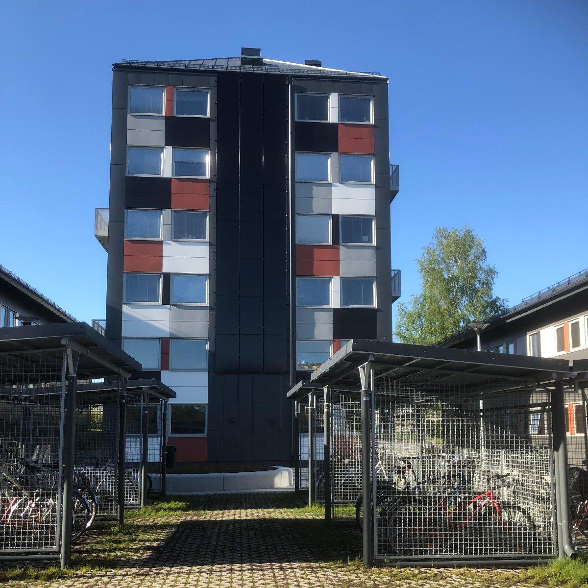 Modernt flervåningshus med färgglada paneler, omgivet av cykelställ under metallskydd och en gräsyta. Klarblå himmel i bakgrunden.