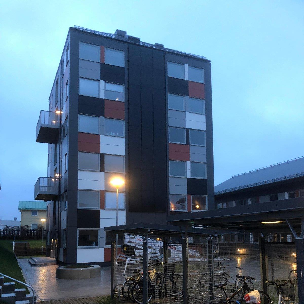 Femvåningshus med modern fasad med rektangulära fönster och paneler i olika färger. Cyklar står parkerade under ett täckt ställ i förgrunden. Mulen himmel i bakgrunden.