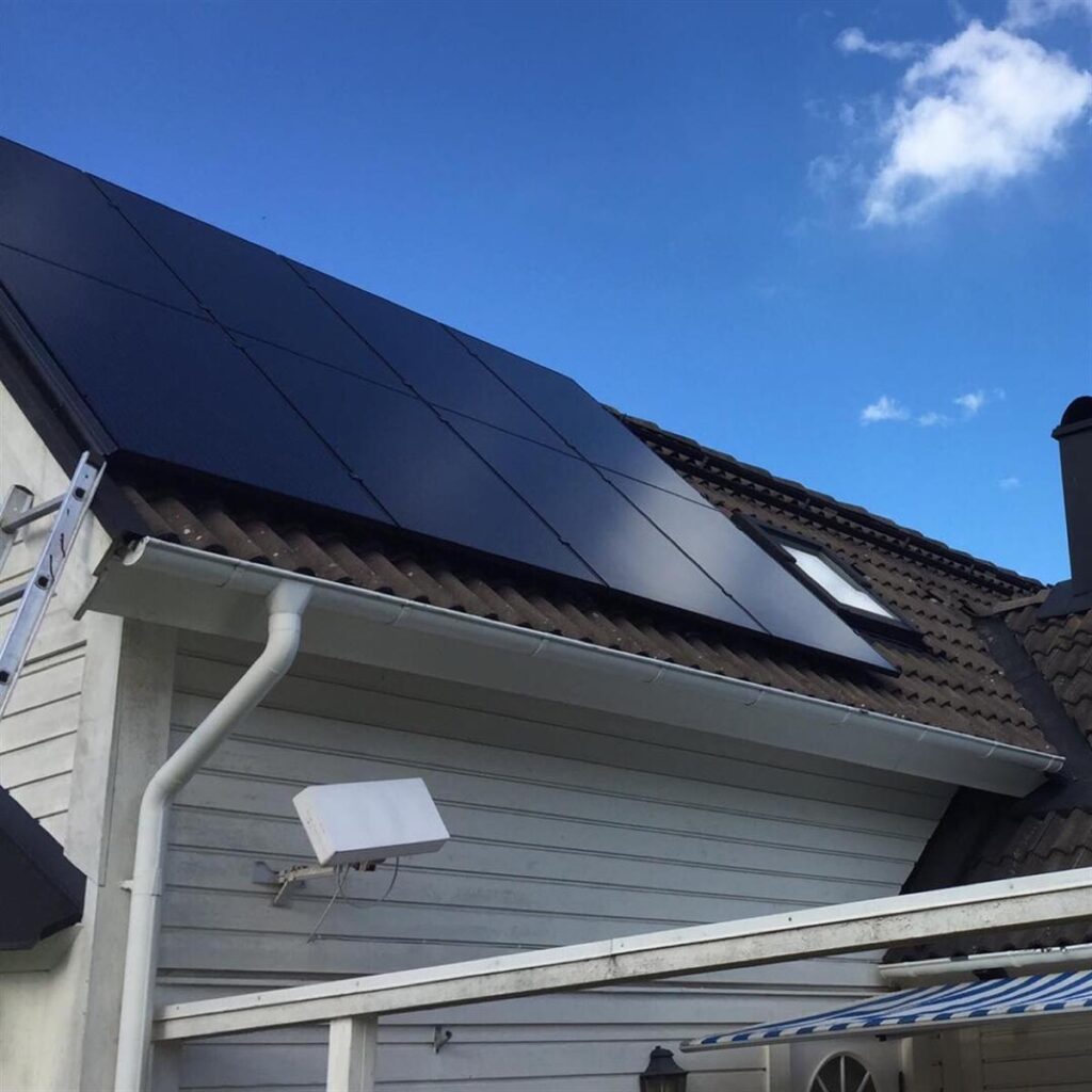 Solpaneler installerade på en sluttande tak av ett vitt hus under en blå himmel med moln. En stege lutar mot taket och en parabol är monterad bredvid panelerna.