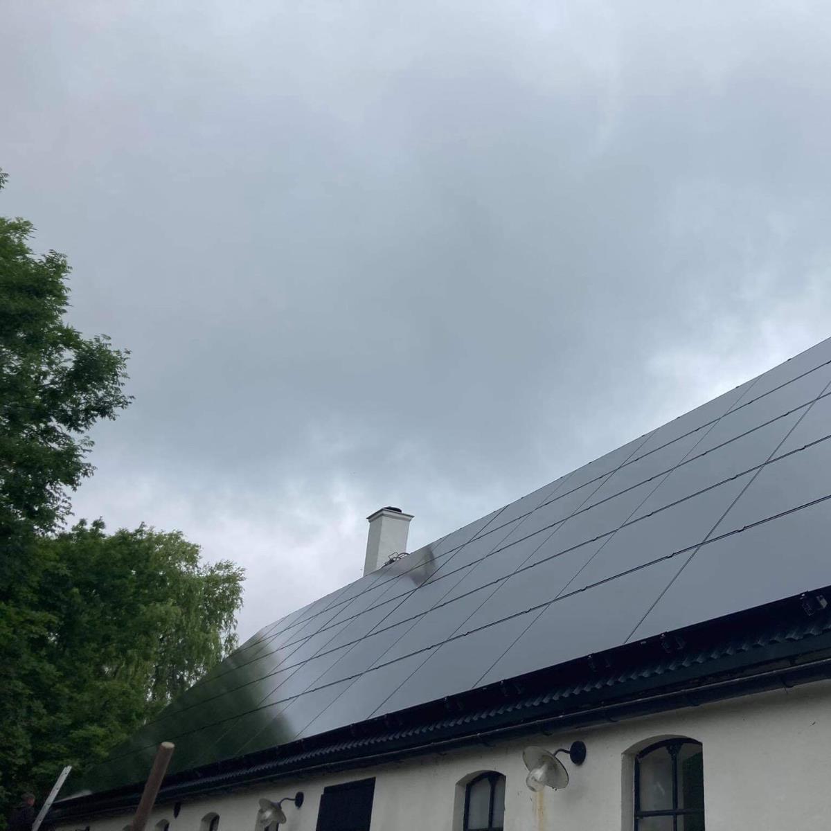 Tak med solpaneler under en molnig himmel, omgiven av grönska.